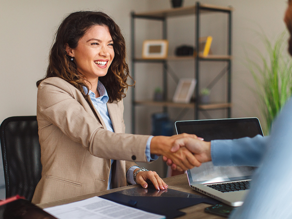 Professional shaking hands with a client across a desk with a laptop and documents nearby